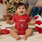 Baby wearing a red Christmas onesie Personalized My 1st Christmas baby bodysuit featuring custom baby character, name, and festive holiday decorations with a Santa Claus toy, sitting on the floor with a decorated Christmas tree in the background.