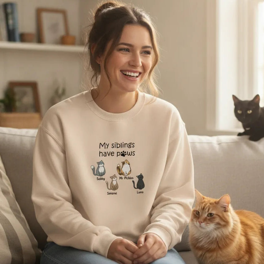 Woman wearing Embroidered My Siblings Have Paws Cats Lightweight Classic Unisex Sweatshirt in Sand, sitting on a couch with cats in a cozy living room.