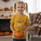 Young girl in a yellow Personalized Little Pumpkin Halloween kids T-shirt with custom child character, name, and pumpkin background holding a small pumpkin in a decorated living room.